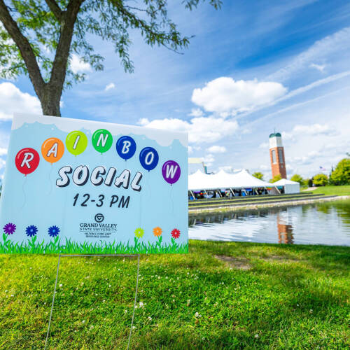 A rainbow social yard sign fills the front view while Zumberge pond, the clocktower, and the Rainbow Social large white tent are visible in the background. The grass is a vibrant green and the blue sky has beautiful fluffy clouds